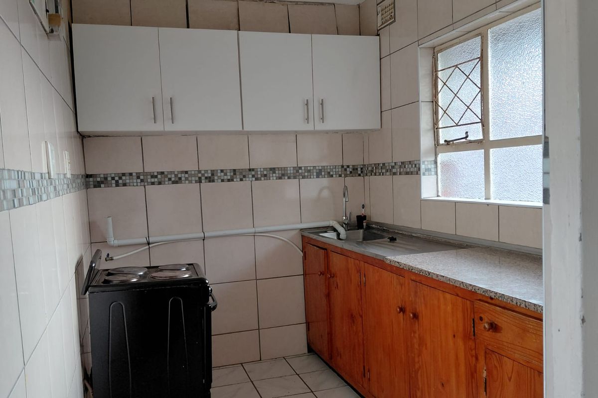 A compact kitchen featuring a mix of wooden and white cabinetry, tiled walls, and a stainless steel sink with exposed surface-mounted plumbing.