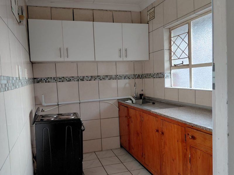 A compact kitchen featuring a mix of wooden and white cabinetry, tiled walls, and a stainless steel sink with exposed surface-mounted plumbing.