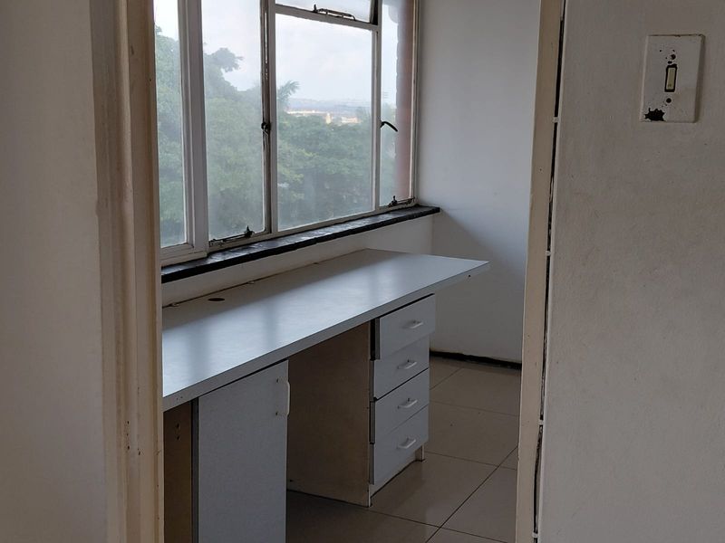 A small, empty office space featuring a built-in desk, tiled flooring, and large metal-framed windows with an exterior view.