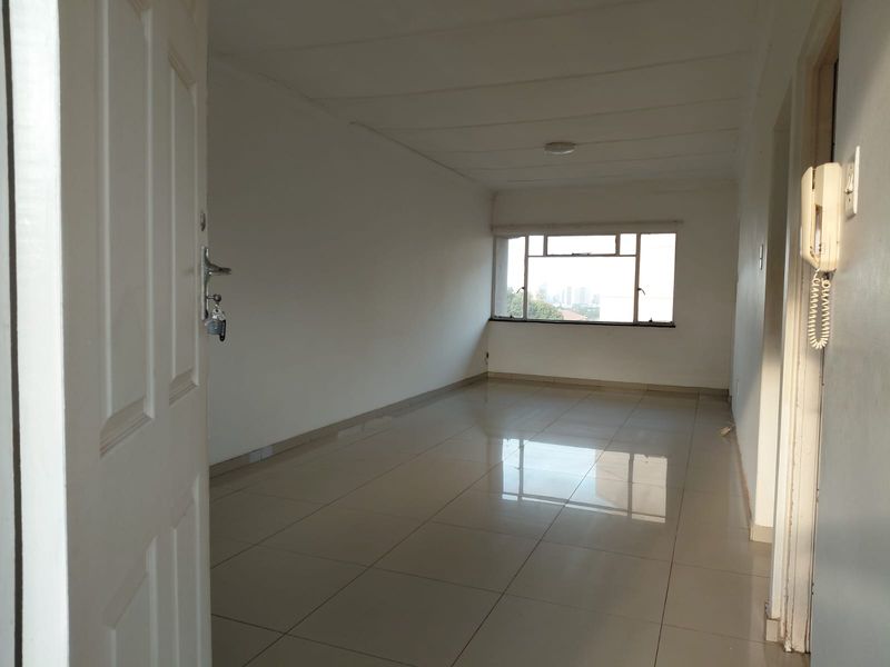 An empty, open-plan living area featuring neutral tiled flooring, white walls, and a large window providing natural light.