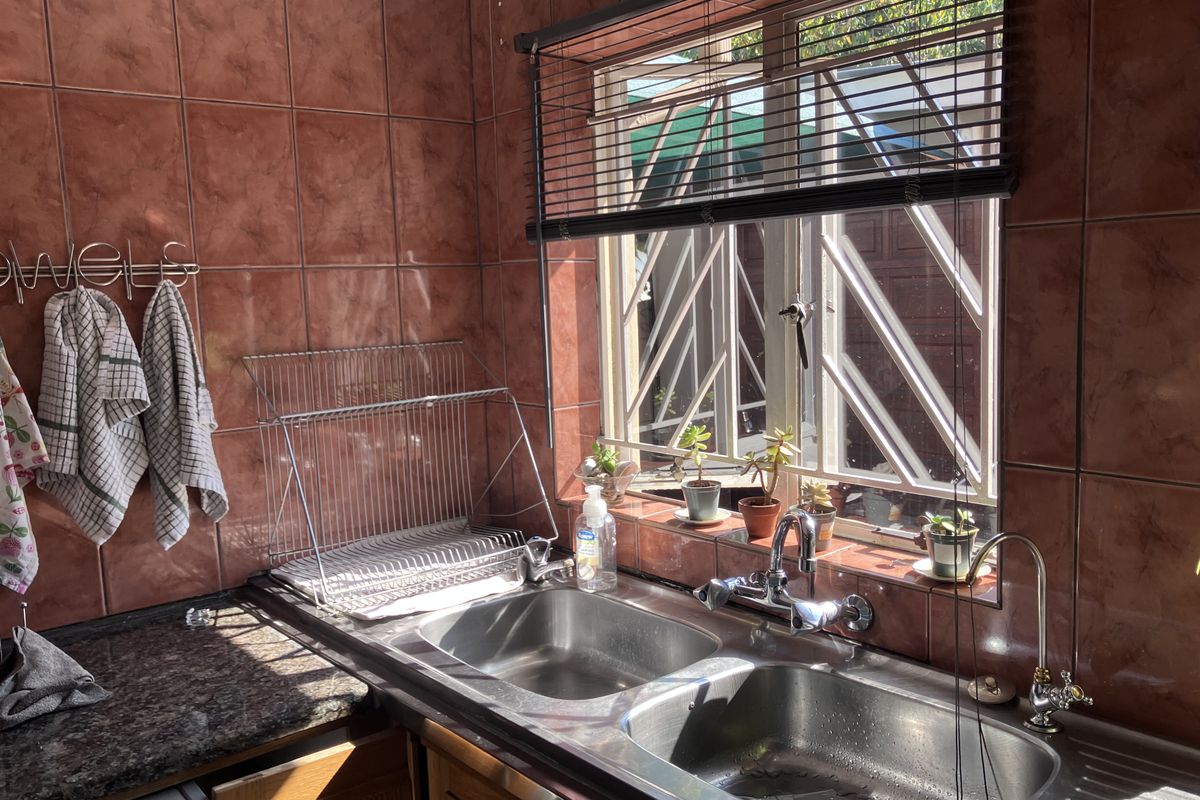 A functional kitchen sink area featuring a double stainless steel basin, granite countertops, and dated wall tiling with security bars on the window.