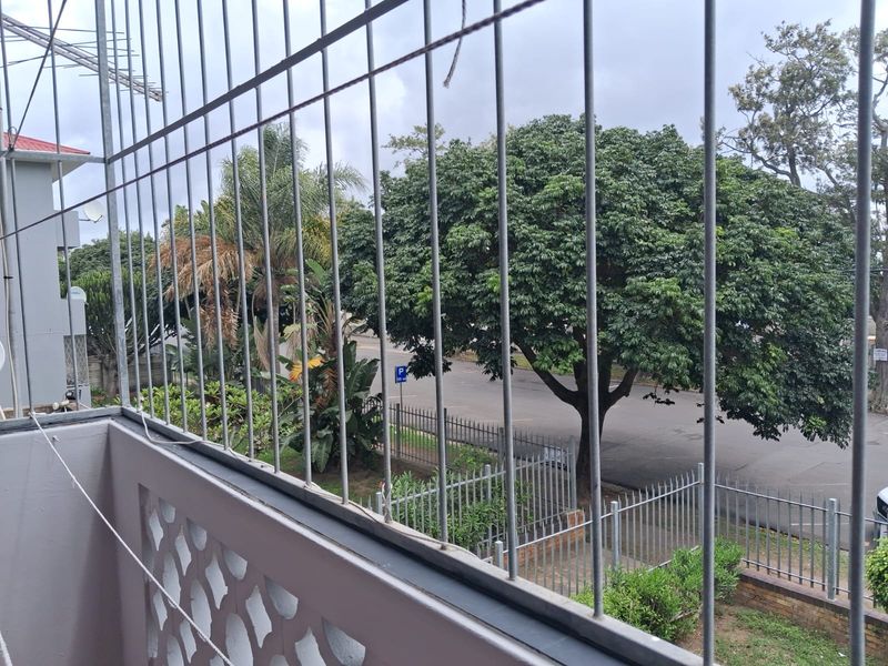 A balcony area featuring decorative concrete balustrades and security bars, overlooking a street with mature trees.