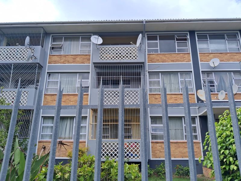 Front elevation of a multi-story apartment building featuring brick and painted concrete facade, private balconies with decorative screens, and perimeter security fencing.