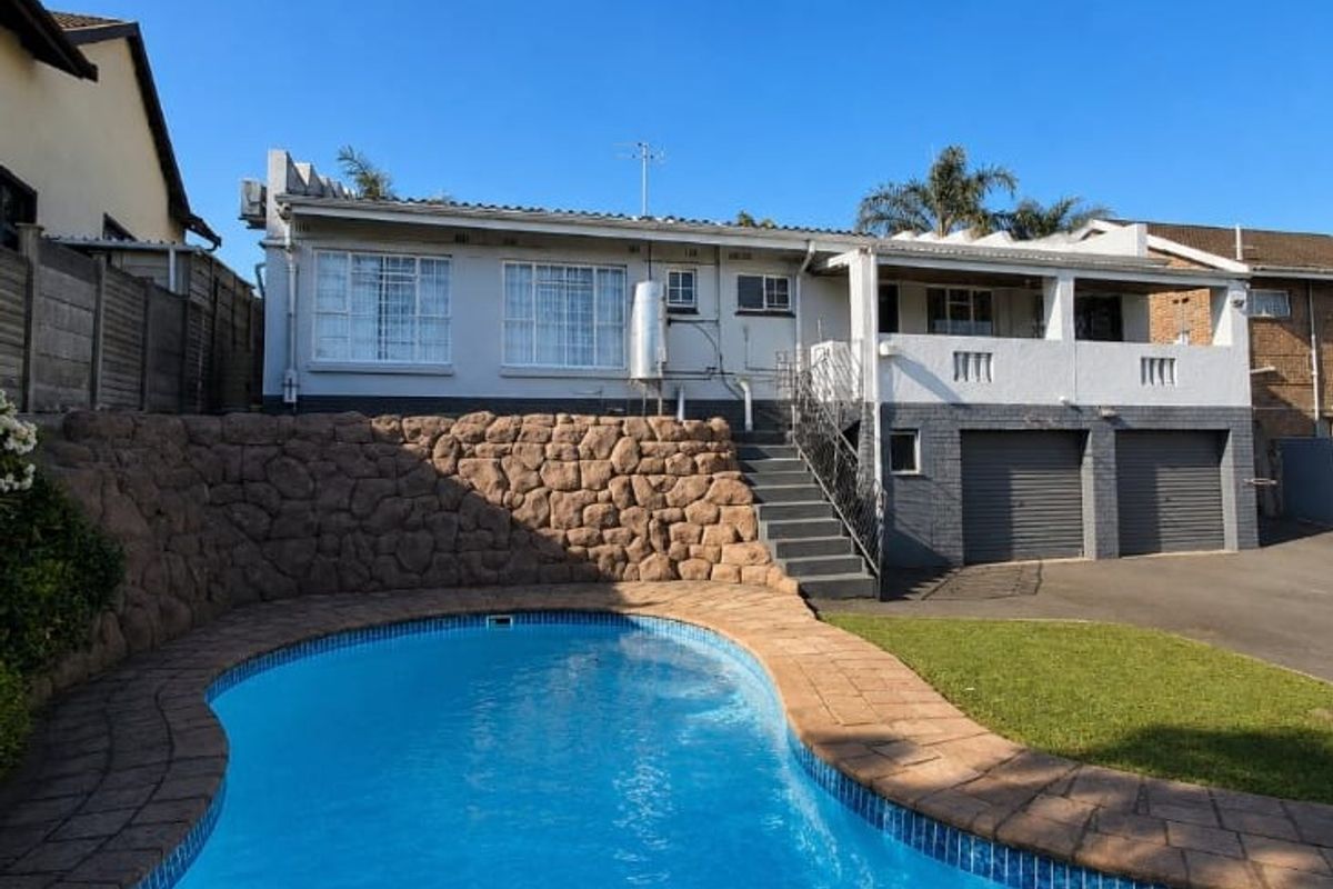 Rear exterior view featuring a swimming pool, stone retaining wall, covered patio, and double garage with paved driveway access.