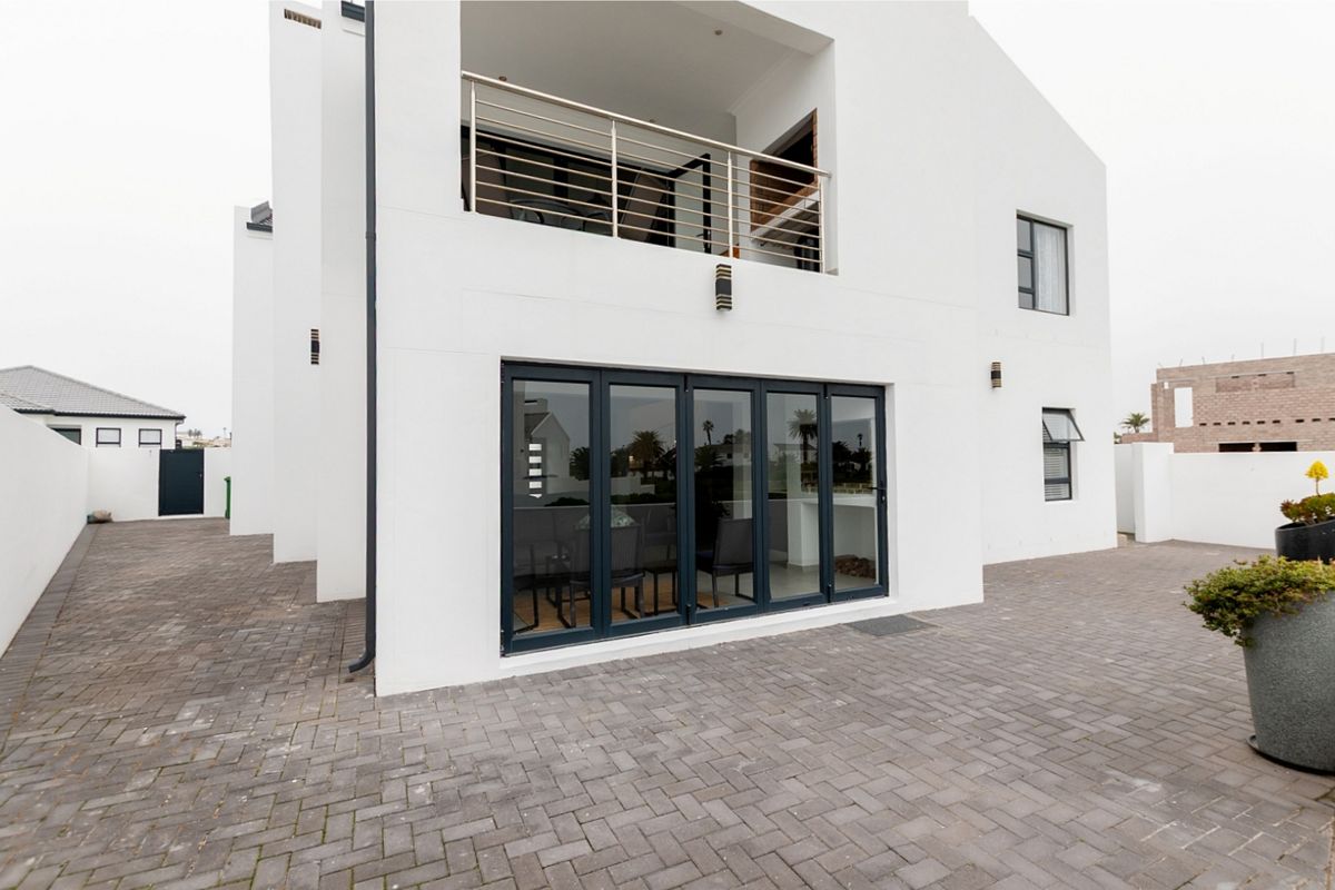 Rear view of a modern two-story house featuring a paved patio area, large dark-framed bi-fold doors, and an upper-level balcony with stainless steel railings.