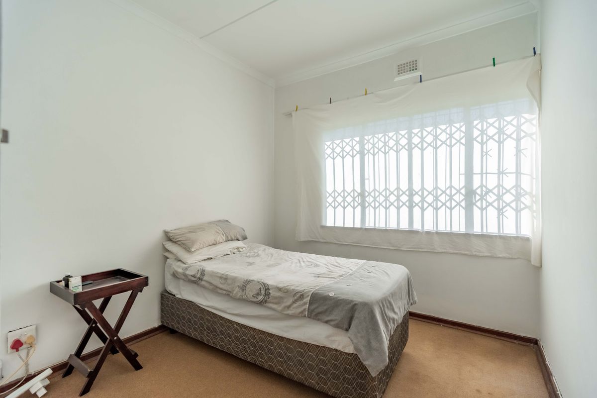 A simple bedroom featuring neutral walls, carpet flooring, and a window fitted with a security gate.