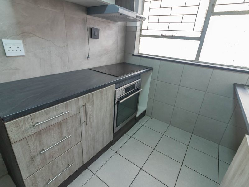 A compact, modern kitchen featuring wood-look cabinetry, dark laminate countertops, built-in oven and hob, and tiled finishes throughout.