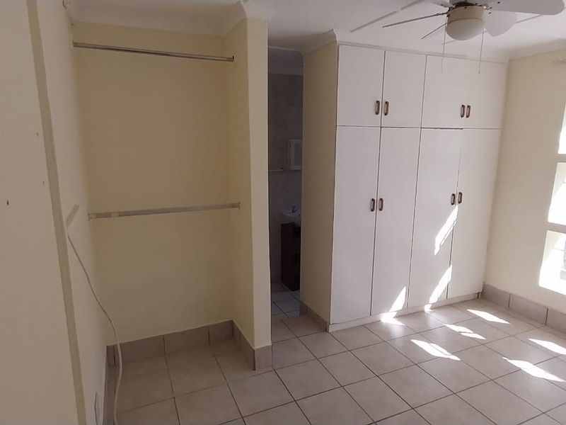 A bright, empty bedroom featuring tiled flooring, built-in cupboards, a ceiling fan, and large windows with security bars.