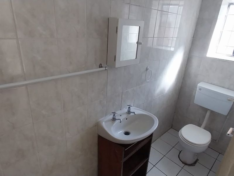 A functional bathroom featuring ceramic wall and floor tiles, a pedestal washbasin, a toilet, and a window with security bars.