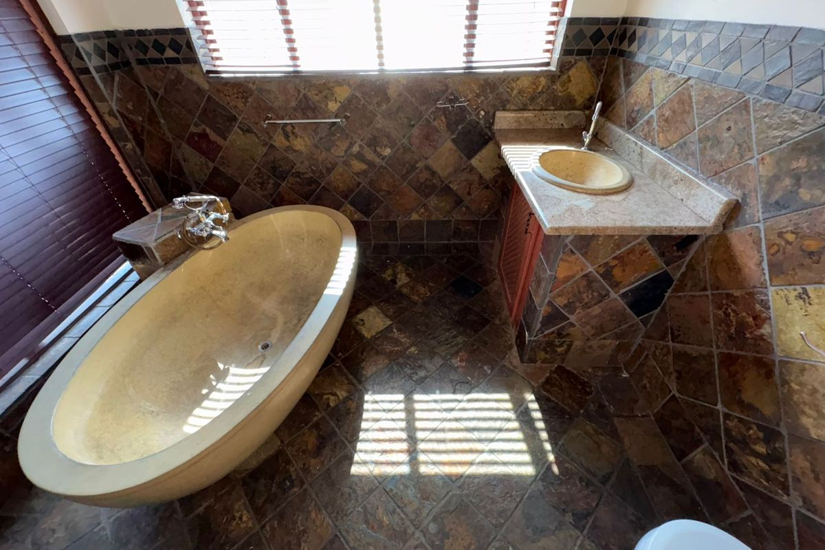 A bathroom featuring a freestanding bathtub, vanity basin, and extensive stone tile wall and floor finishes with natural light from venetian blinds.