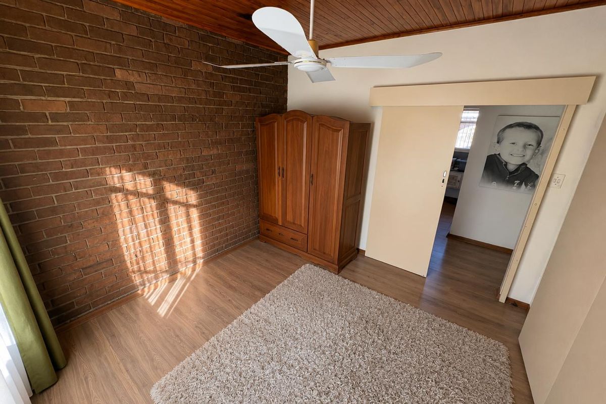 A bedroom featuring an exposed brick feature wall, timber ceiling, ceiling fan, and laminate flooring, with a sliding door leading to an adjacent room.