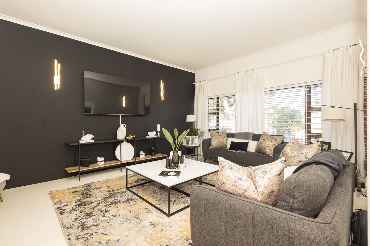 A modern living room featuring a bold black accent wall, large windows with venetian blinds, and contemporary wall-mounted lighting fixtures.