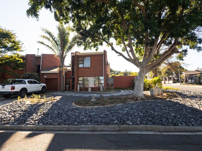 Front view of a brick townhouse featuring a paved driveway, integrated garage, and a landscaped front garden with mature trees.