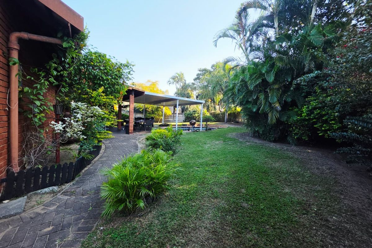 A spacious rear garden featuring a paved walkway, a covered patio area, and a swimming pool surrounded by established landscaping.