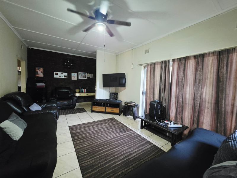 A spacious living room featuring tiled flooring, a ceiling fan, and a dark brick feature wall with access to the outdoors via sliding glass doors.