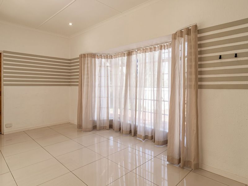 A bright, empty living room featuring large bay windows, neutral tiled flooring, and a wooden door, offering a clean canvas for interior design.