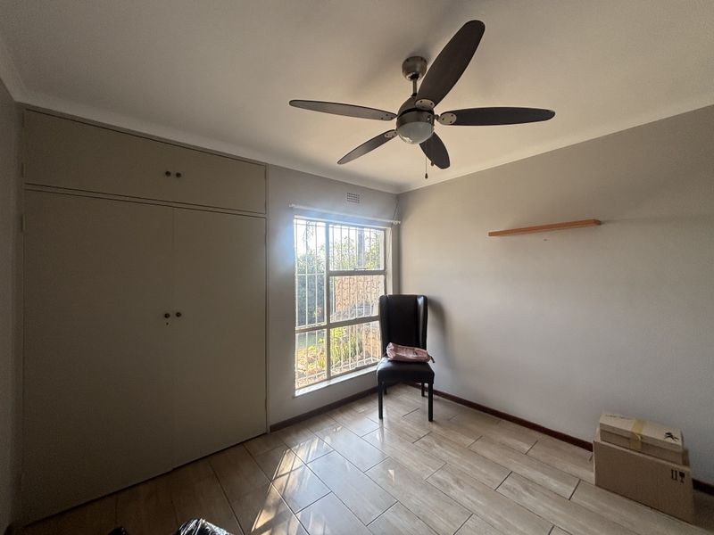 A bright bedroom featuring tiled flooring, built-in cupboards, and a ceiling fan, with security bars installed on the window.
