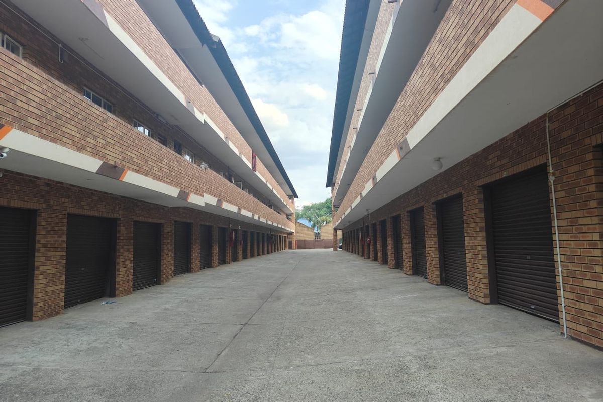 Exterior view of an apartment complex featuring a central concrete driveway flanked by rows of face brick units with individual roller shutter garage doors.