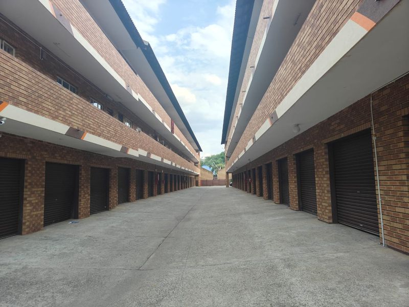 Exterior view of an apartment complex featuring a central concrete driveway flanked by rows of face brick units with individual roller shutter garage doors.