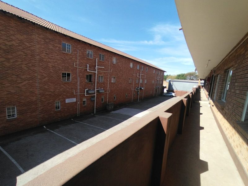 An exterior view of the apartment complex showing the rear brick facade, a concrete walkway with balustrade, and the paved parking area below.