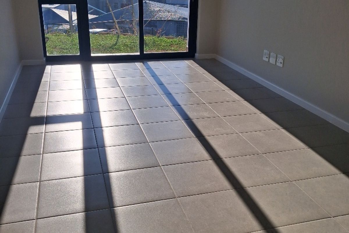 An empty, sunlit living area featuring neutral floor tiles, modern skirting boards, and a large sliding glass door with views of the surrounding landscape.