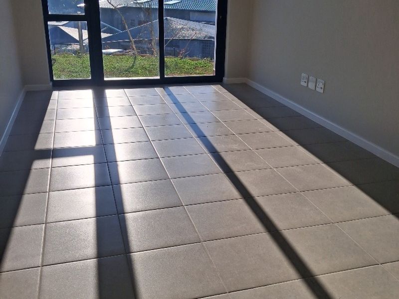 An empty, sunlit living area featuring neutral floor tiles, modern skirting boards, and a large sliding glass door with views of the surrounding landscape.
