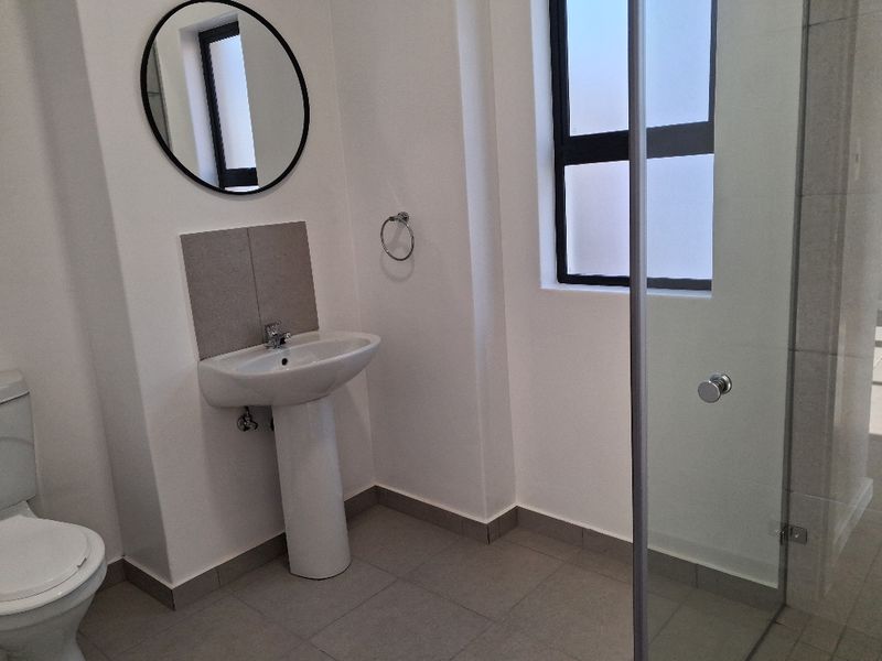 A clean, modern bathroom featuring a pedestal basin, toilet, and a glass-enclosed shower with neutral floor tiling.