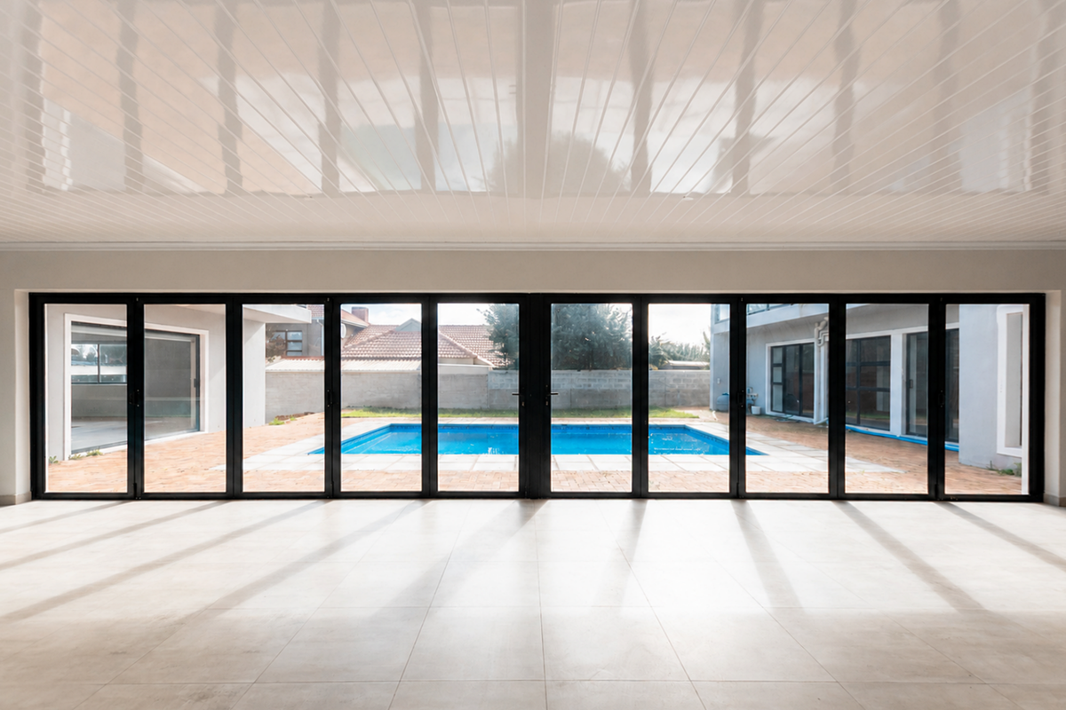 Spacious, empty living area featuring large format floor tiles, a glossy ceiling, and expansive black aluminium stacker doors opening onto a swimming pool.