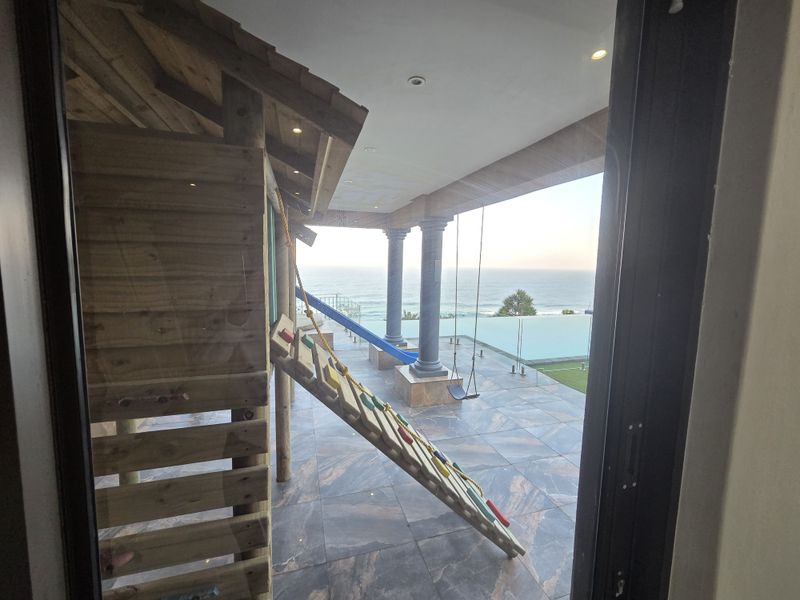 Spacious covered patio featuring high-end tiled flooring, stone columns, and a glass balustrade overlooking an infinity pool with ocean views.
