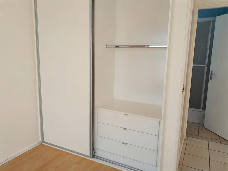 A bedroom featuring a built-in sliding door wardrobe with internal shelving and drawers, complemented by laminate flooring and an adjacent tiled hallway.