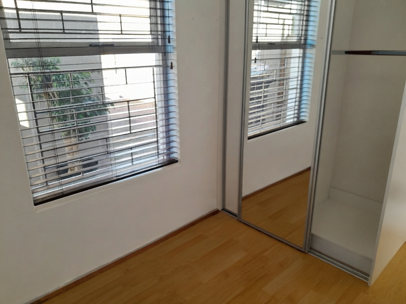 A bright, empty bedroom featuring laminate flooring, a built-in mirrored wardrobe, and venetian blinds on the windows.
