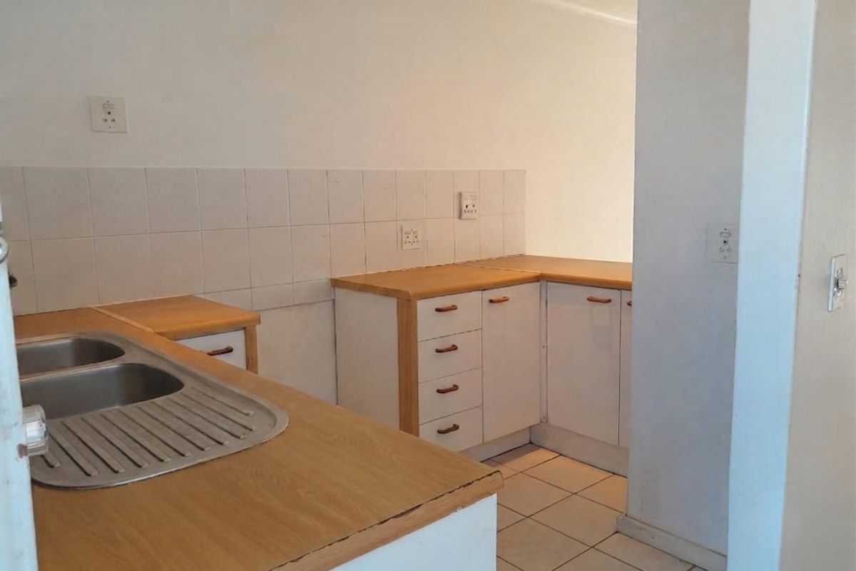 A compact, empty kitchen featuring laminate countertops, a double bowl stainless steel sink, tiled backsplash, and white cabinetry with wood trim.