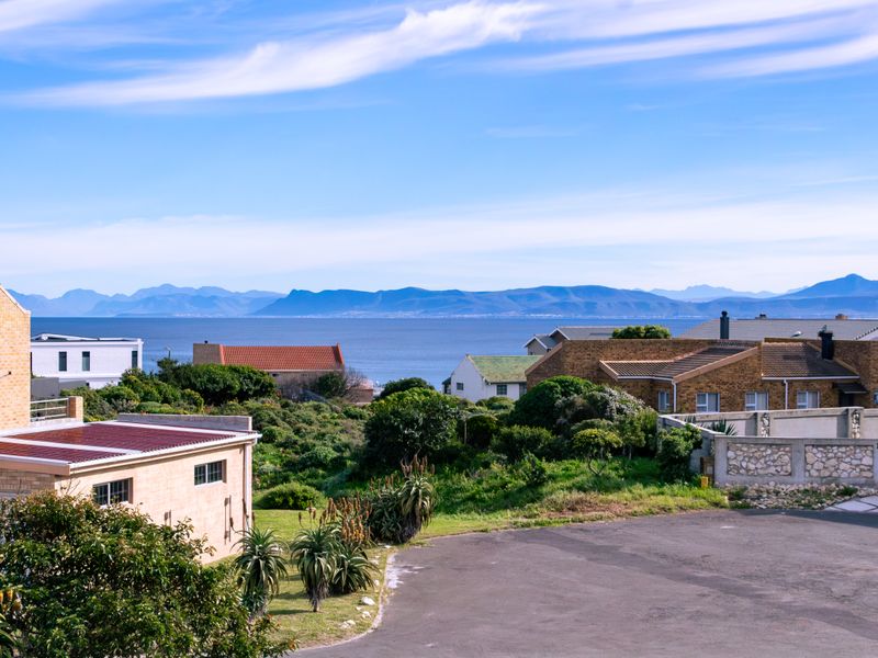 Elevated coastal view overlooking residential rooftops, lush greenery, and the ocean with distant mountain ranges on the horizon.