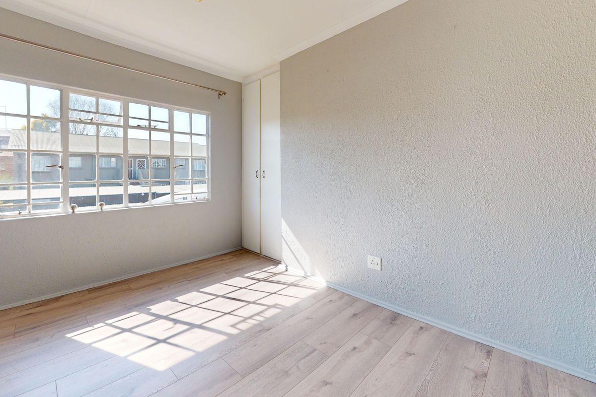 A bright, empty bedroom featuring light-toned laminate flooring, a built-in wardrobe, and a large multi-pane window allowing for natural light.
