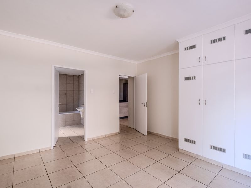 A spacious, empty bedroom featuring neutral tiled flooring, built-in white cupboards, and direct access to an en-suite bathroom.