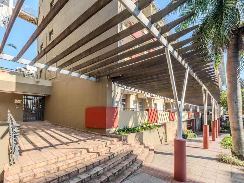 Exterior view of the apartment building entrance featuring a large timber pergola, paved walkway, and concrete stairs leading to the main security gate.