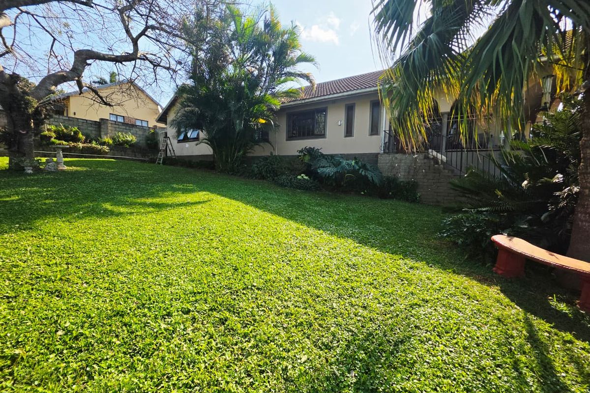 A spacious rear garden featuring a well-maintained lawn, mature trees, and a raised patio area with brick retaining walls and metal railings.