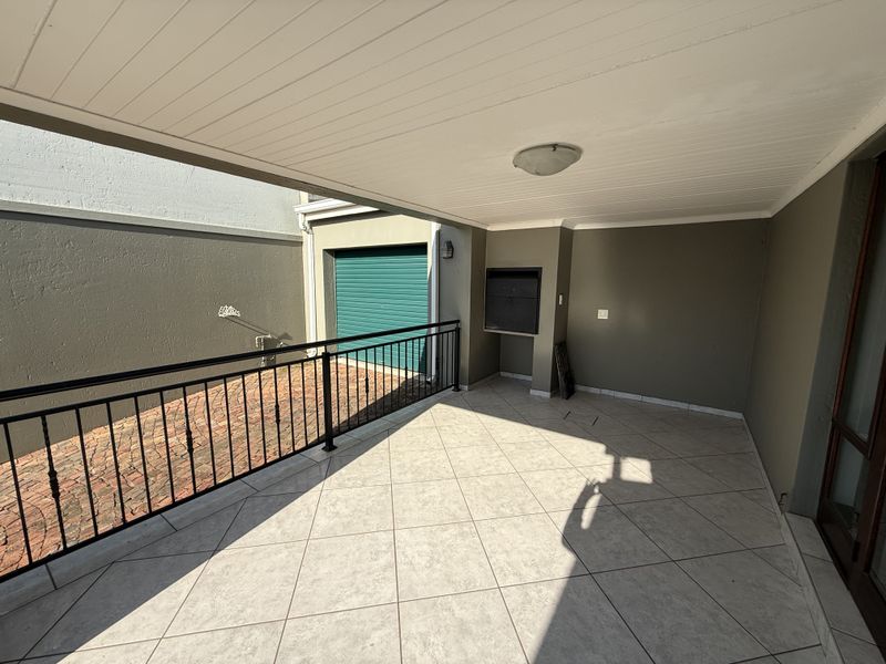Covered patio area featuring tiled flooring, a built-in braai, and a metal balustrade, overlooking a paved courtyard and garage entrance.