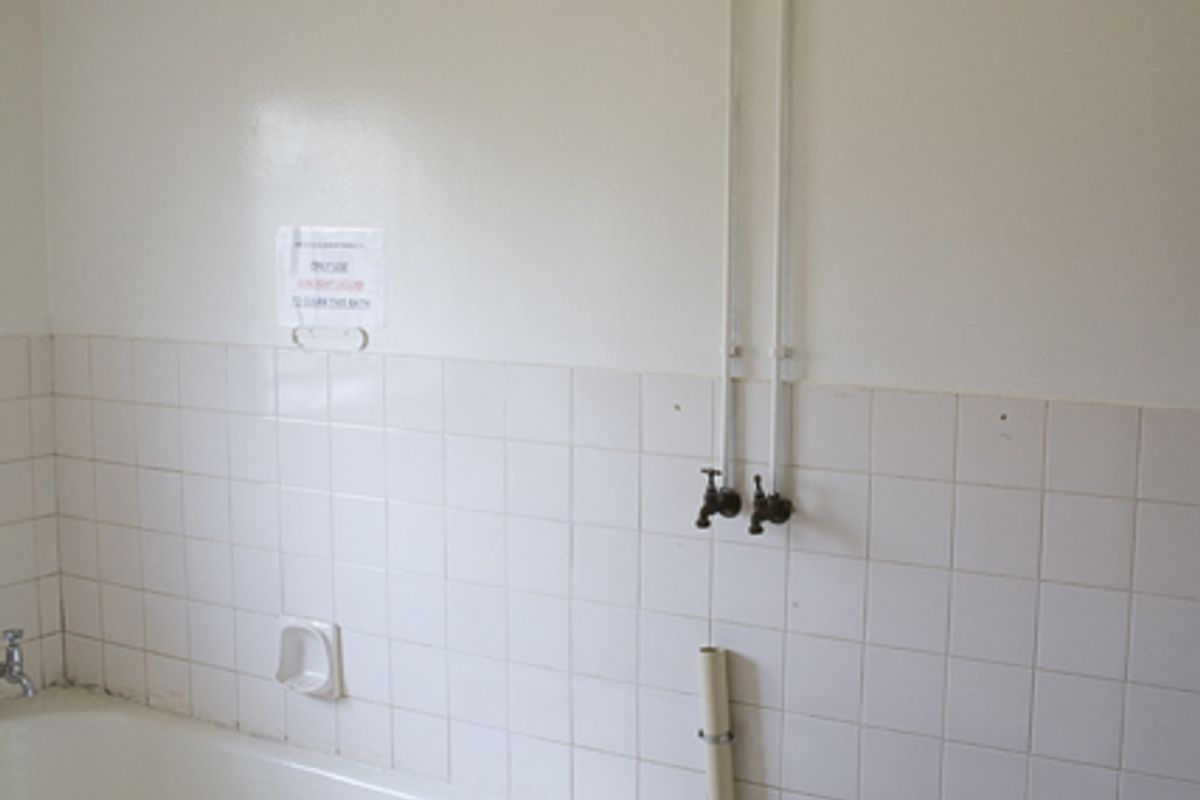 A dated bathroom featuring a tiled bathtub, exposed plumbing fixtures, and white wall tiles. The space is vacant and requires cosmetic updates.