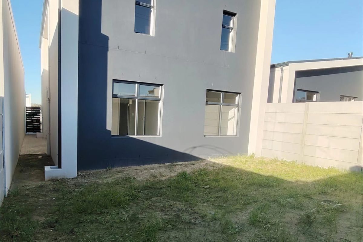 Rear view of a modern two-story house featuring a painted facade, aluminium windows, and an unlandscaped garden area enclosed by a precast concrete wall.