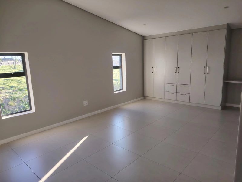 A spacious, modern bedroom featuring neutral tiled flooring, built-in cupboards, and large windows providing ample natural light.