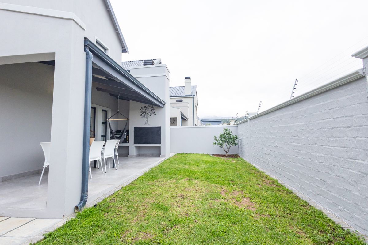 A neat rear garden featuring a covered patio with a built-in braai, tiled flooring, and a secure perimeter wall with electric fencing.