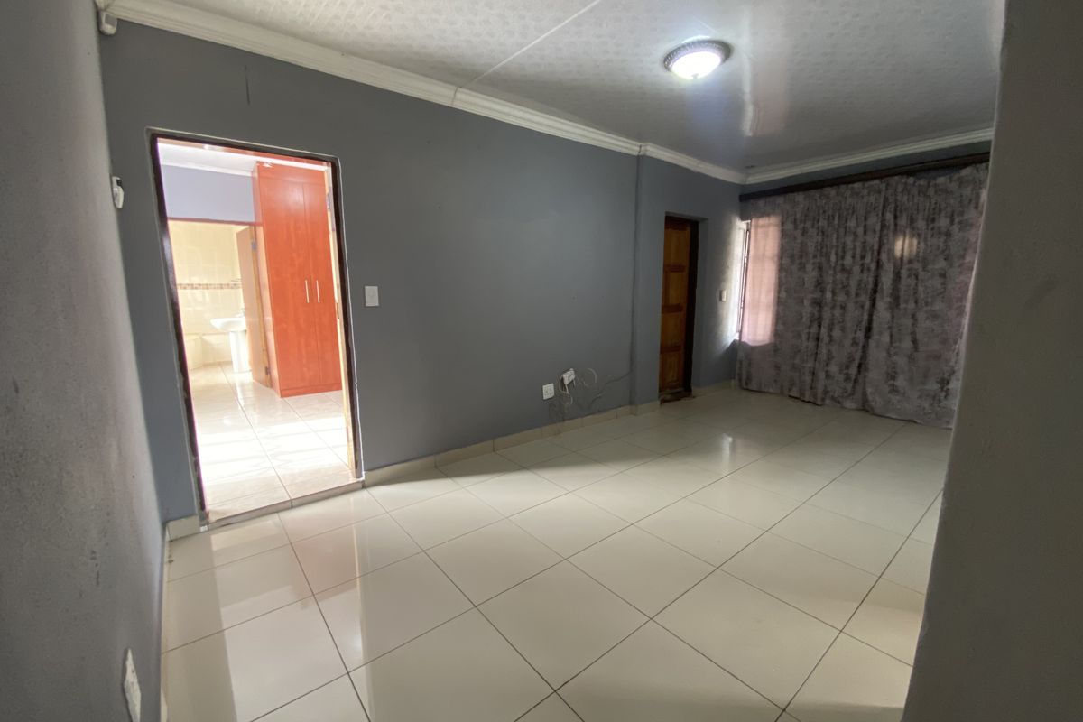 A spacious, empty living area featuring neutral grey walls, tiled flooring, and a view into an adjacent room with built-in cabinetry.