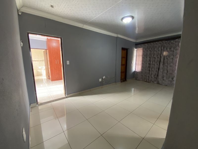 A spacious, empty living area featuring neutral grey walls, tiled flooring, and a view into an adjacent room with built-in cabinetry.