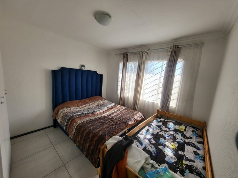 A functional bedroom featuring tiled flooring, large windows with security bars, and neutral wall finishes, providing a practical space for rest.