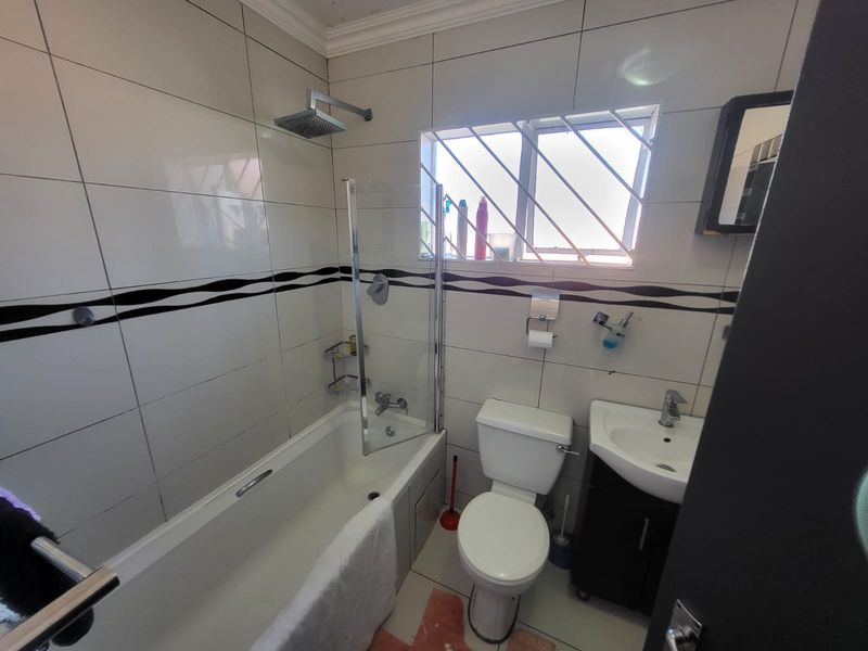 A functional bathroom featuring a bathtub with an overhead shower, glass screen, vanity basin, and tiled walls with decorative border accents.