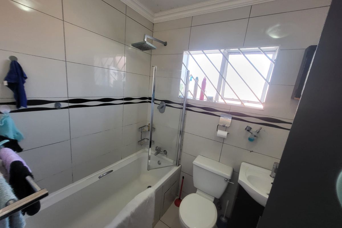 A functional bathroom featuring a bathtub with a glass shower screen, toilet, and vanity basin, finished with tiled walls and security bars on the window.