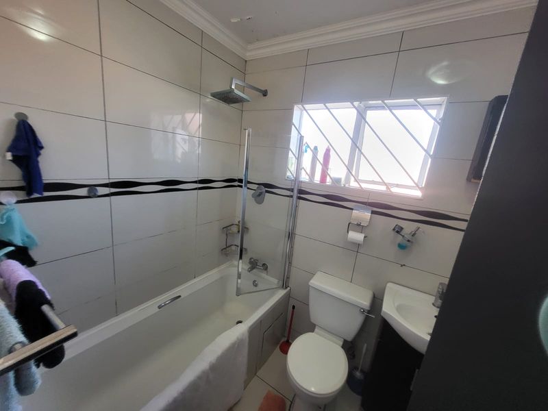 A functional bathroom featuring a bathtub with a glass shower screen, toilet, and vanity basin, finished with tiled walls and security bars on the window.
