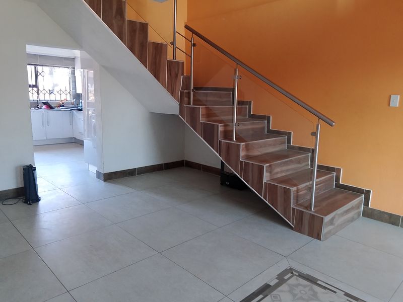 Open plan living area featuring a modern tiled staircase with stainless steel and glass balustrade, leading towards a kitchen area with security gate.