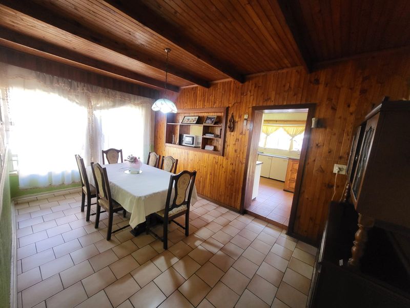 Dining room featuring timber-clad walls, exposed beam ceiling, and ceramic floor tiles, with an adjacent view into the kitchen area.